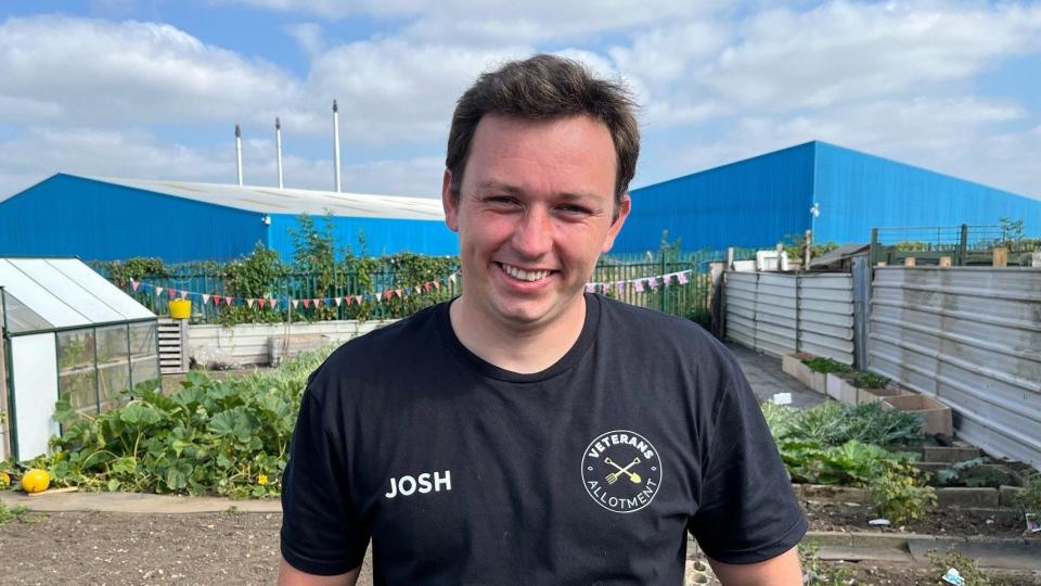 Josh is standing at an allotments wearing a black t-shirt with a veterans allotment logo on. He has short brown hair and is smiling for the camera.