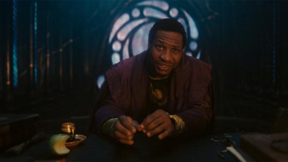 Image from the Marvel T.V. show Loki, season 2 episode 6. A man with short dark hair and robes is sitting at a large desk in a dark room, with a circular window behind him.