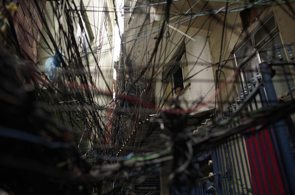 FILE - In this March 20, 2020 file photo, a senior citizen peers from his window through a web of electric wires in the Rocinha slum of Rio de Janeiro, Brazil, amid orders to stay home to contain the spread of the coronavirus. In Rocinha, narrow alleyways reduce airflow around homes packed tightly together; clean water is scarce, sewage often runs in the streets and winding alleys and soaring staircases make it difficult for medical professionals to retrieve an ailing patient in case of emergency. (AP Photo/Silvia Izquierdo)