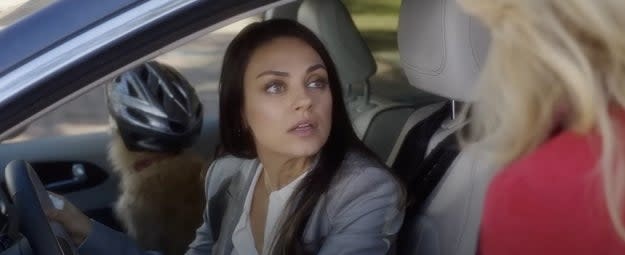 Mila Kunis looks out the window from a car, with a dog wearing a bike helmet in the backseat. An unidentified person in a red outfit stands outside the car