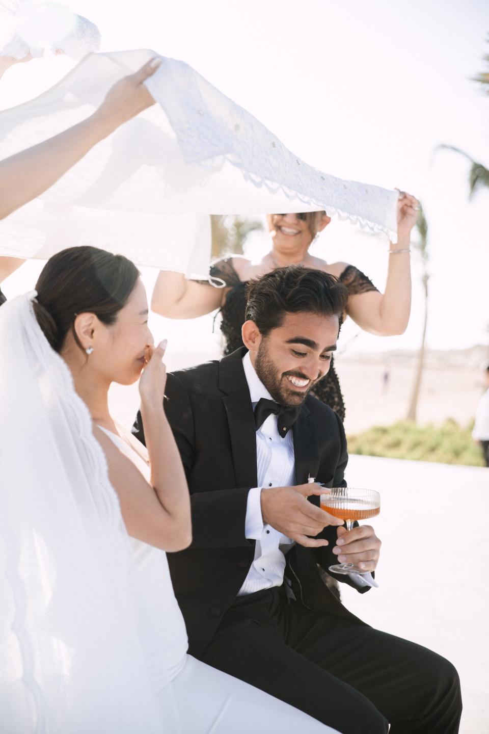 A bride and groom laugh as people hold a veil over their heads.