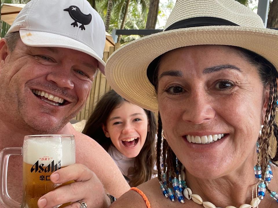 Man holding a beer with woman and young girl, with palm trees in the background.