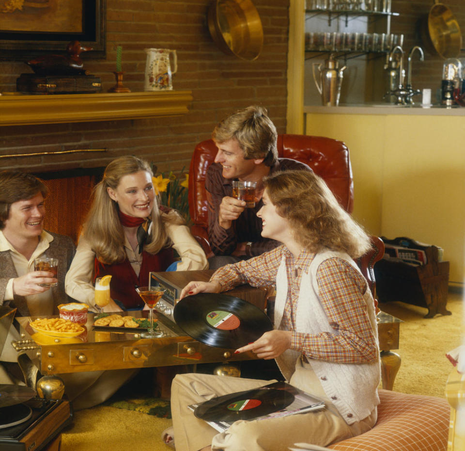 A group of four adults enjoy drinks and snacks around a coffee table, listening to vinyl records in a retro living room