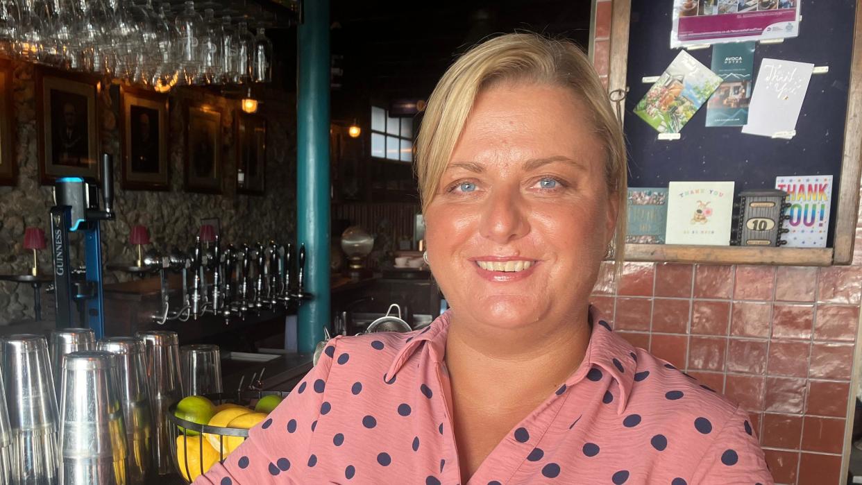 Nicola Stephenson is wearing a pink polkadot linen looking shirt. 

She has blonde hair tied up in a pony tail. Her hair is in a side parting and she has blue eyes. She is smiling at  the camera. 

She is standing in front of a bar stacked with wine glasses and draught beer. Beside the bar there is a fruit bowl and plastic cups. 