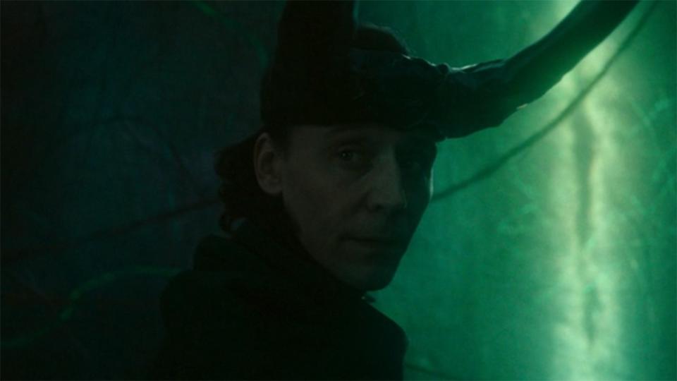 Image from the Marvel T.V. show Loki, season 2 episode 6. Close up of Loki's face, the God of Mischief. He is looking back over his shoulder solemnly as he steps towards a green portal. He is wearing a headpiece with two long, black horns on his head.