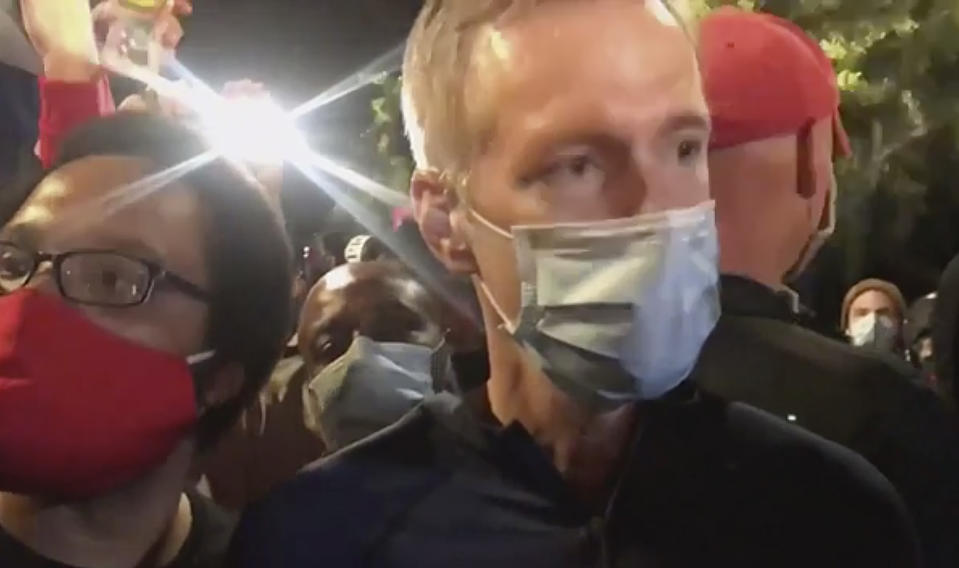 In this image made from video released by Karina Brown, Portland Mayor Ted Wheeler, right, stands at a fence guarding a federal courthouse as tear gas drifts by early July 23, 2020, in Portland Oregon, during another night of protest against the presence of federal agents sent by President Donald Trump to quell unrest in the city. (Karina Brown via AP)