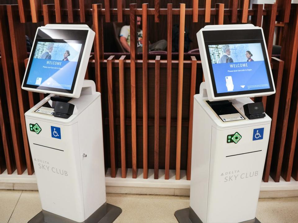 Self check-in stations with blue "welcome" screens and white bodies in front of a wall with wooden panels