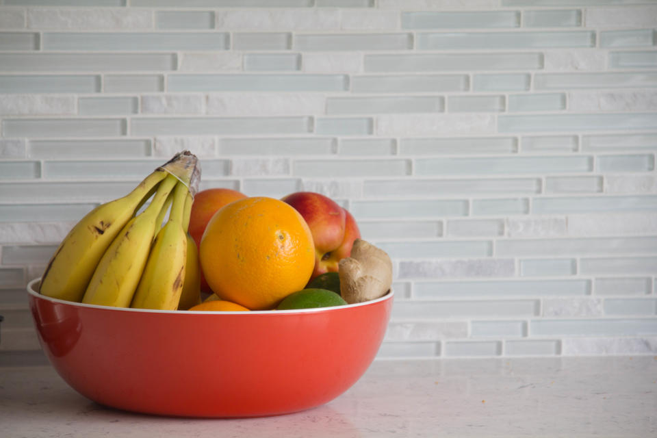 Red bowl with fresh fruit on grey granite kitchen counter with glass tile backsplash.