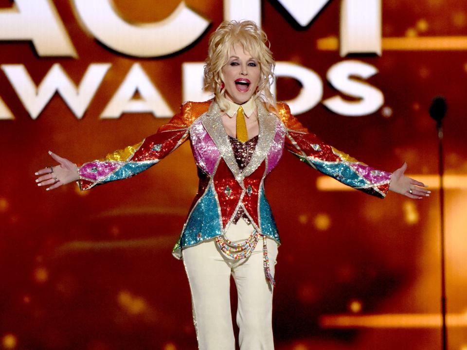 Dolly Parton with her arms open wide wearing a shiny, colorful patchwork blazer and cream-colored pants.