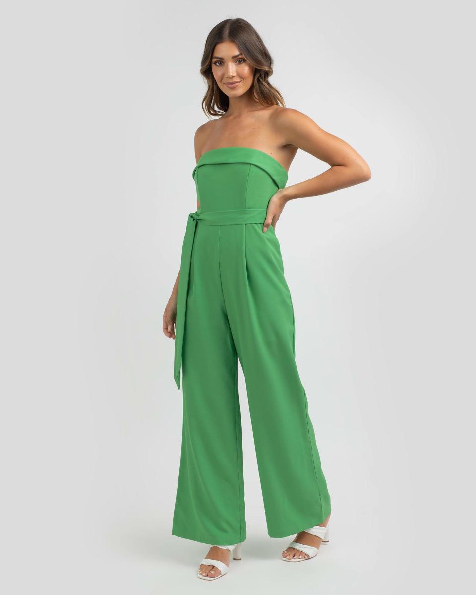 A model with shoulder length brown hair worn loose wears a bright green Winnie & Co Fabbie Jumpsuit, $59.99, with white sandals against a grey background.