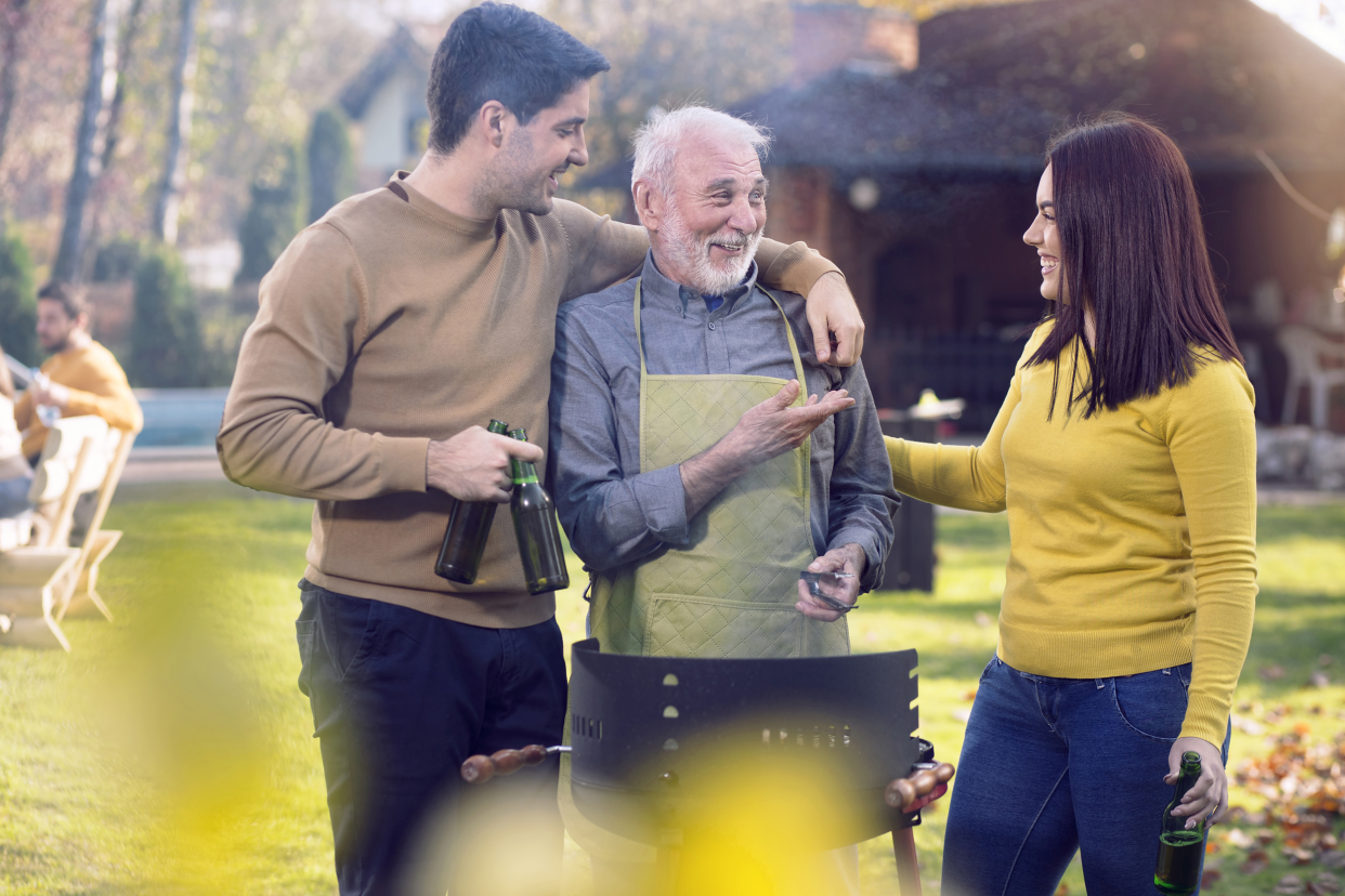 Young man hugging senior man who is at the grill with young woman on side, all are smiling and engaged in light-hearted conversation, outside during a autumn in yard of a large home, with a blurred background of trees and another home