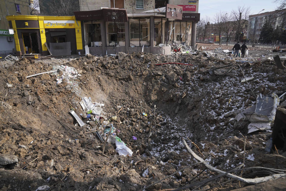 People walk past a crater from the explosion in Mira Avenue (Avenue of Peace) in Mariupol, Ukraine, Sunday, March 13, 2022. The surrounded southern city of Mariupol, where the war has produced some of the greatest human suffering, remained cut off despite earlier talks on creating aid or evacuation convoys. (AP Photo/Evgeniy Maloletka)