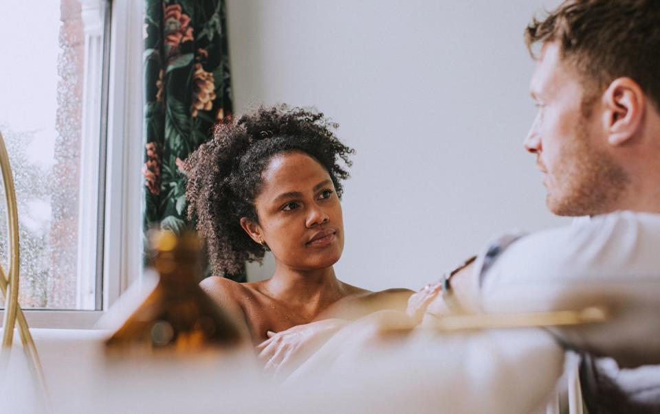 serene image of a woman bathing in a roll top bath her partner kneels by the side of the tub, and rests his arms on the side they look at each other as they have a conversation they look relaxed and comfortable with each other