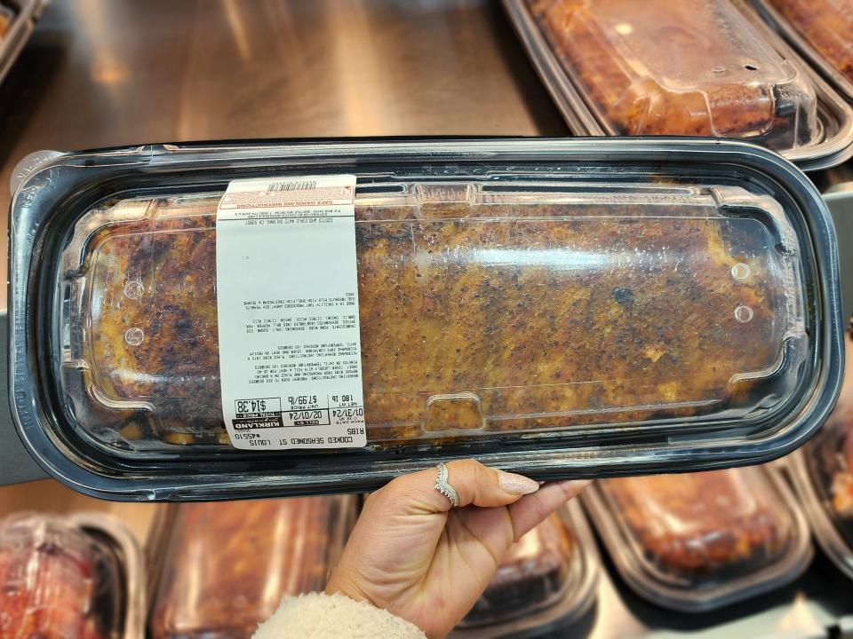 Hand holding Kirkland Signature St. Louis ribs in front of rotisserie chickens and other meat at Costco