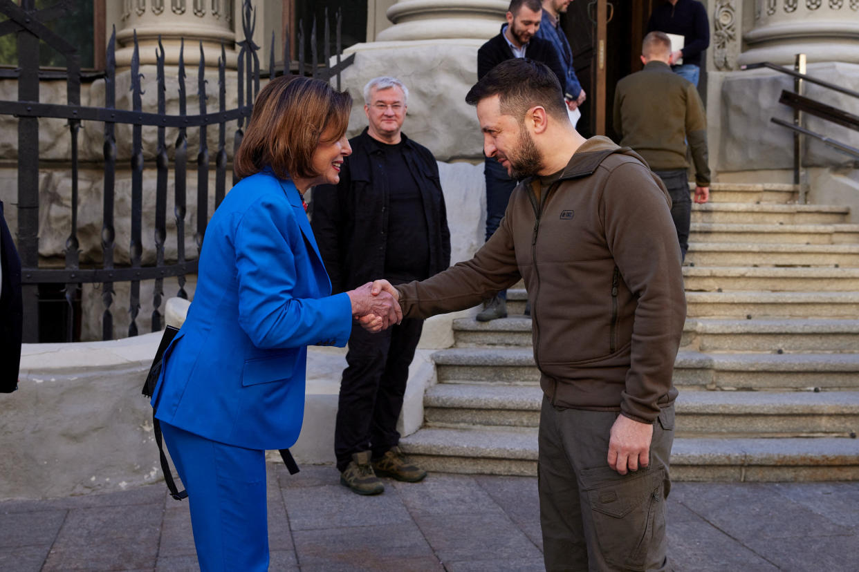 Ukrainian President Volodymyr Zelensky shakes hands with Nancy Pelosi on a sidewalk near a building's entryway in front of a few onlookers.