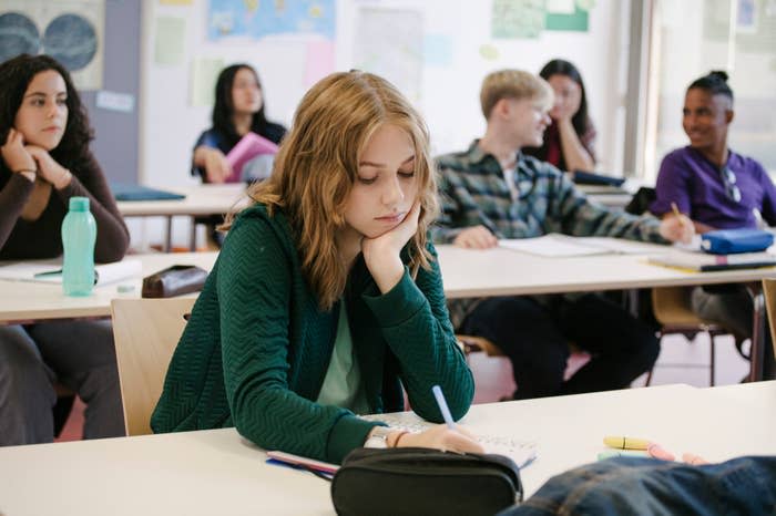 A student looks bored, resting her cheek on her hand, while others listen to a lecture in a classroom setting