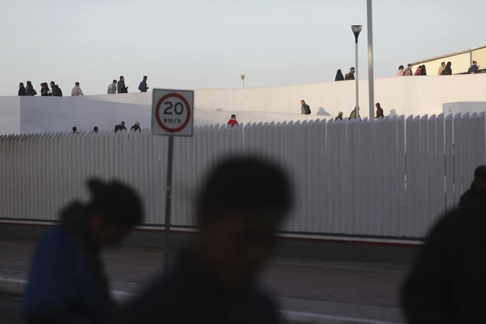 Pedestrians cross the Chaparral border to the U.S. as migrants, foreground, begin to gather nearby, in Tijuana, Mexico, Friday, Nov. 23, 2018. The mayor of Tijuana has declared a humanitarian crisis in his border city and says that he has asked the United Nations for aid to deal with the approximately 5,000 Central American migrants who have arrived in the city. (AP Photo/Rodrigo Abd)