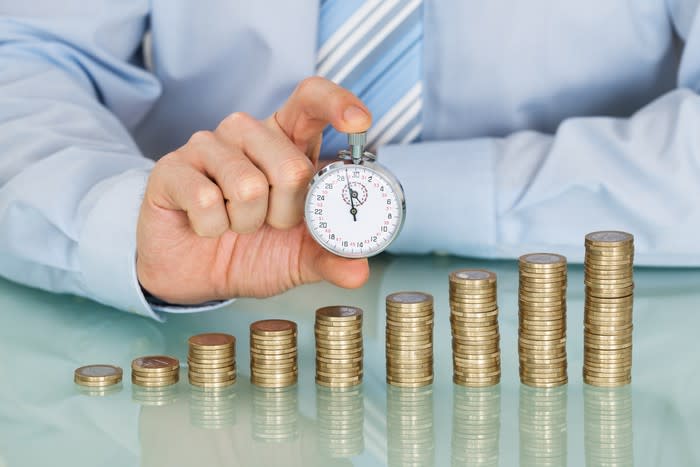 A businessman holding a stopwatch behind an ascending pile of coins.