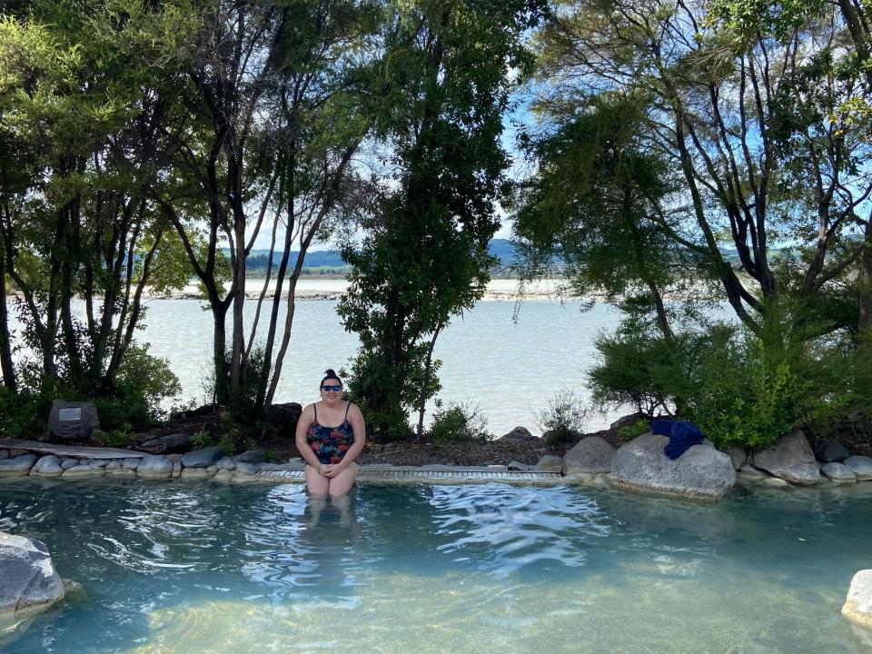 Author Riana Ang-Canning sitting on a stone edge in a Polynesian spa in front of a body of water in New Zealand