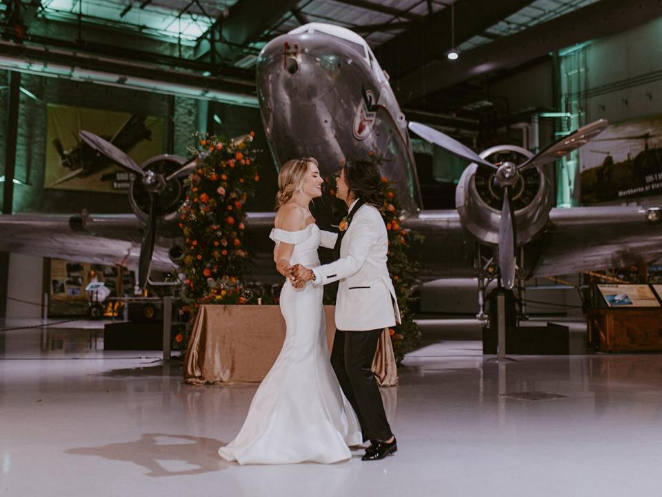 Two brides dance in front of an airplane.