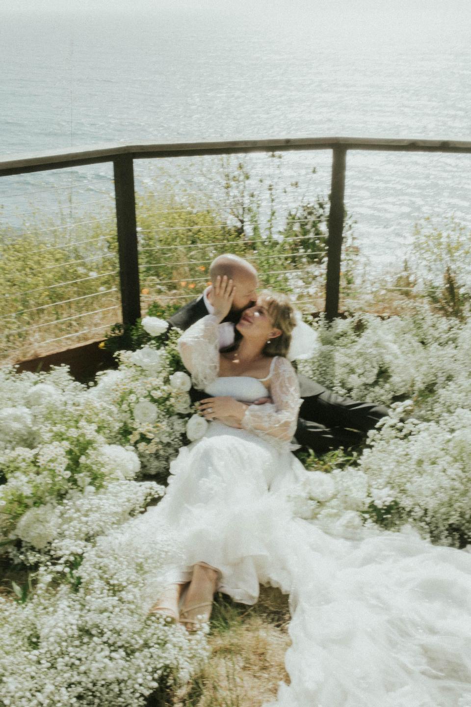 A bride and groom embrace and look at each other while sitting in a field of flowers.