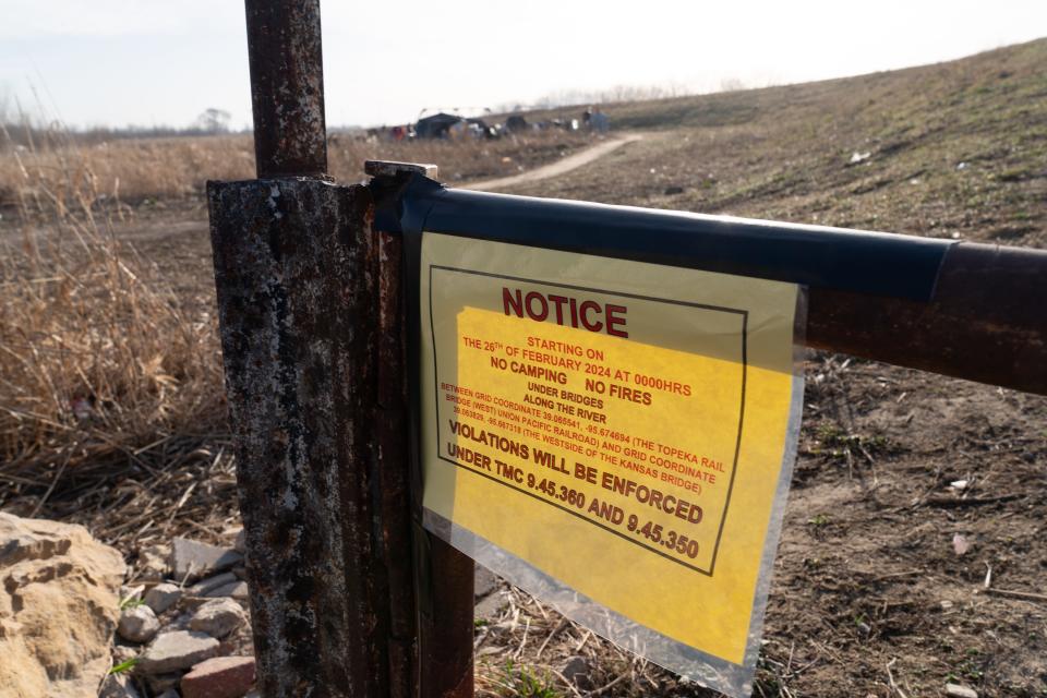 A notice sign is posted just east of the Kansas Avenue Bridge and north of the Kansas River stating violation enforcement for camping and fires within the area homeless have been living.