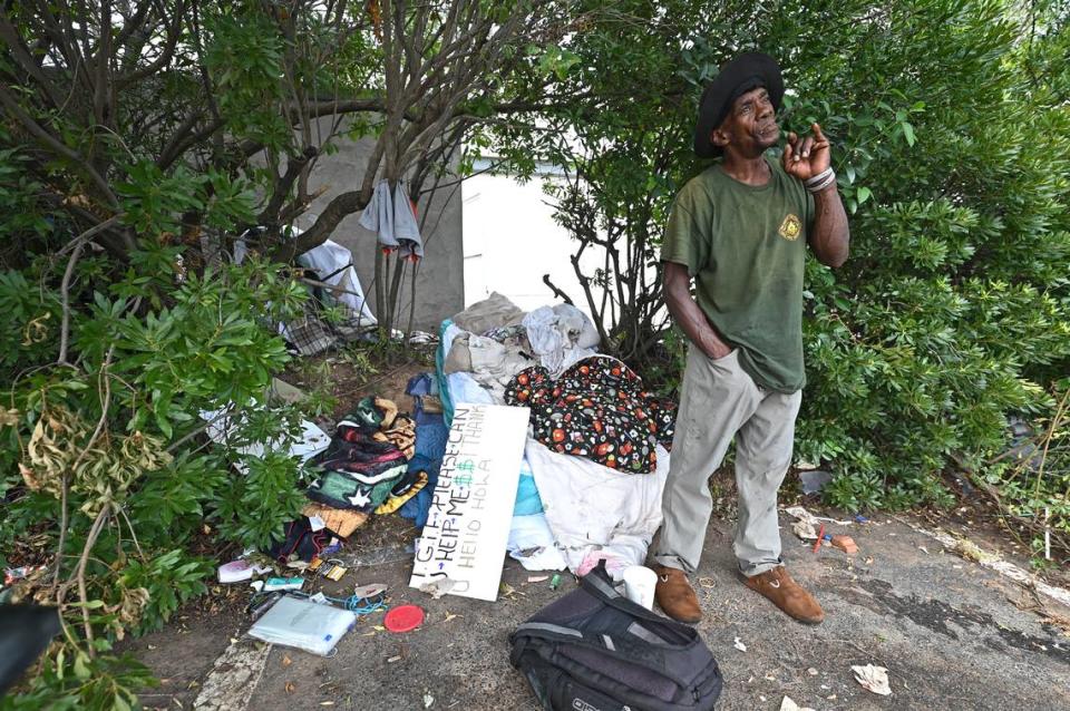 Kenny puffs on a cigar as he stands next to his collection of belongings in the bushes next to an old Boost Mobile store.