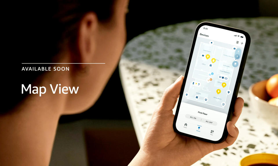 Amazon product photo showcasing Map View. View from over a person's right shoulder as they hold a smartphone. The phone's screen shows a digital floor plan with icons representing smart devices around it.