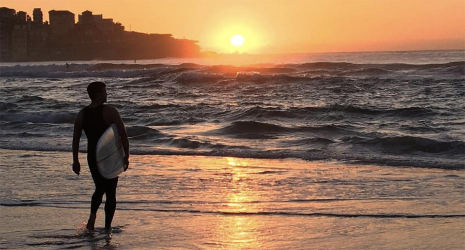 A surfer stands in the shallows at sunrise on Bondi Beach.