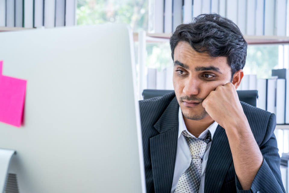 A man wearing a suit and tie sits at a desk, resting his face on his hand, looking at a computer screen with a bored expression