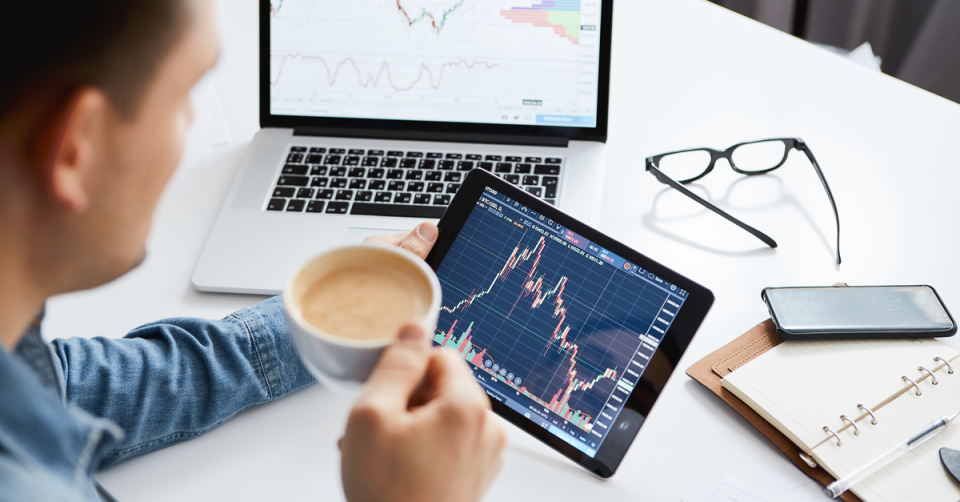 a man drinks a cup of coffee while looking at the stock market on his computer tablet