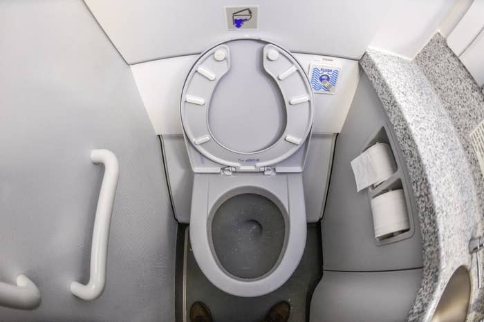 Airplane lavatory with an open toilet lid, gray handle, and two rolls of toilet paper on a shelf