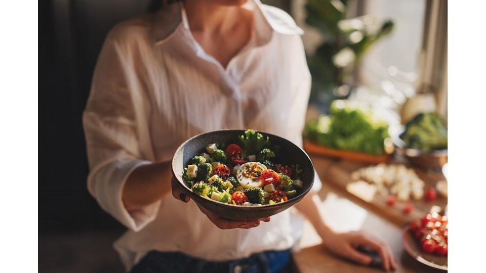 Healthy dinner or lunch. Woman in t-shirt and jeans standing and holding vegan superbowl or Buddha bowl with hummus, vegetable, salad, beans, couscous and avocado and smoothie in hands, square crop