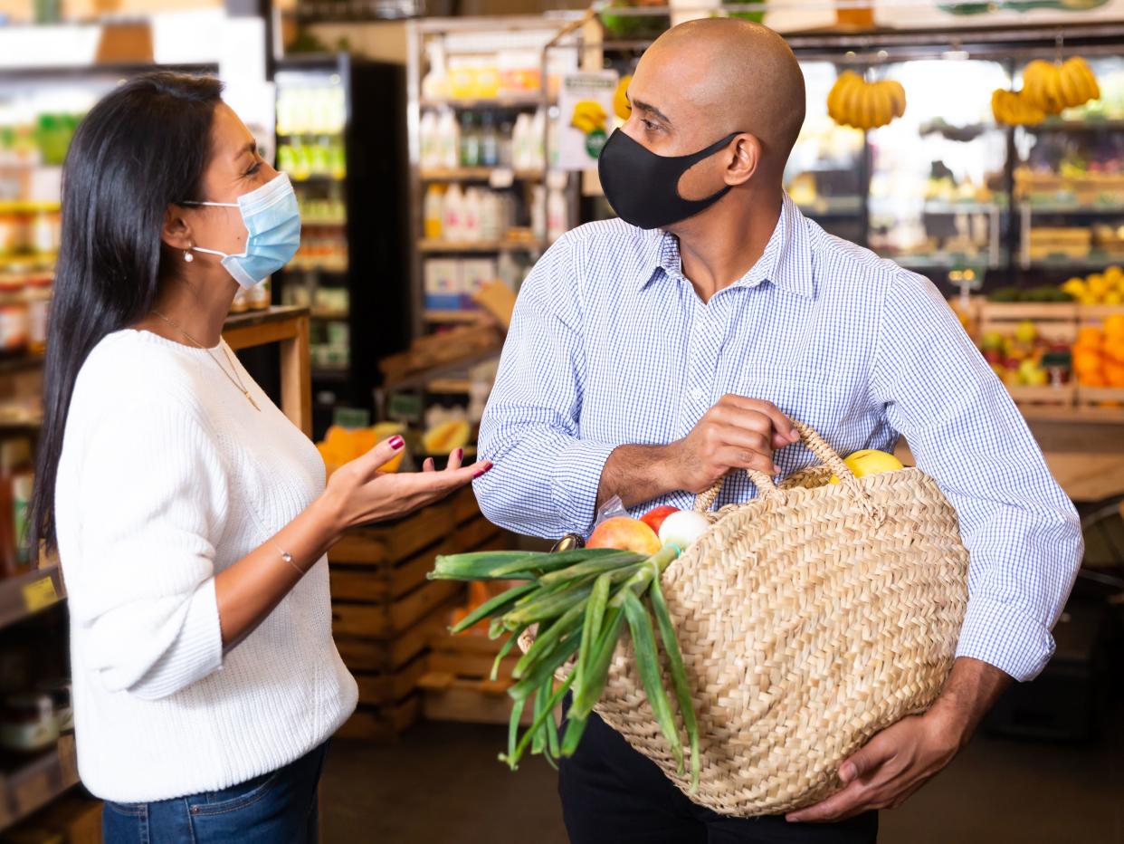 Smiling Latino wearing protective mask to prevent viral infection standing with purchases in grocery store, friendly talking to woman