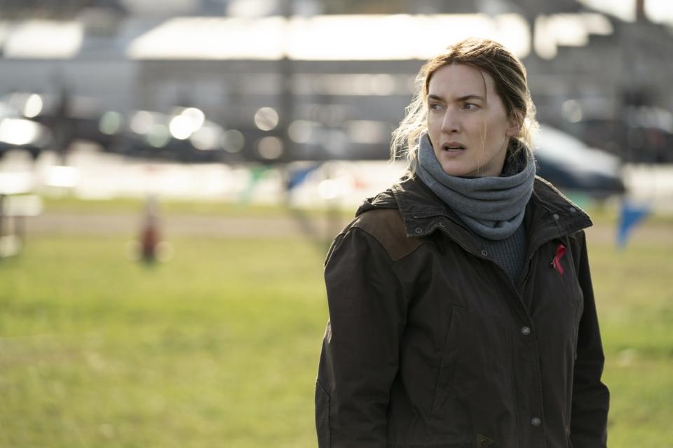 Kate Winslet stands outdoors on a stretch of grass in a winter jacket and gray muffler.