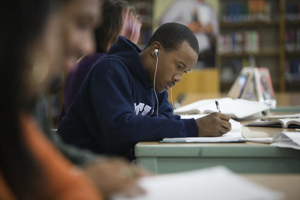 A Black male teenager studies at a library while listening to music through his earphones.