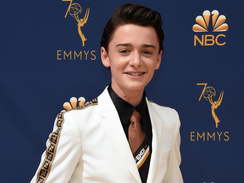 noah schnapp wearing a white suit with gold detailing stripes on the sleeves, along with a brown tie and black shirt. he's standing in front of a blue background with the 70th emmys logo and the nbc logo