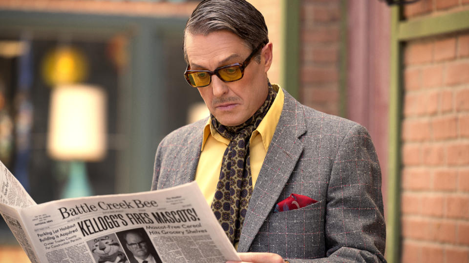 Hugh Grant reads a newspaper in the alley with a look of discomfort in Unfrosted.