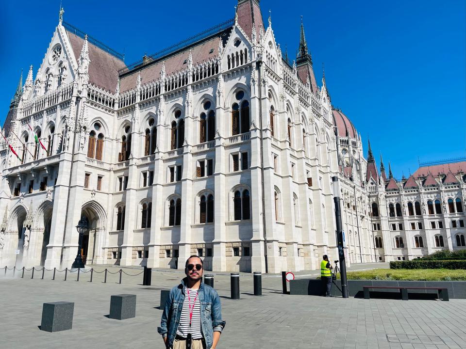 A man with a striped shirt, jean jacket, and sunglasses poses in front of the parliament building in Budapest.