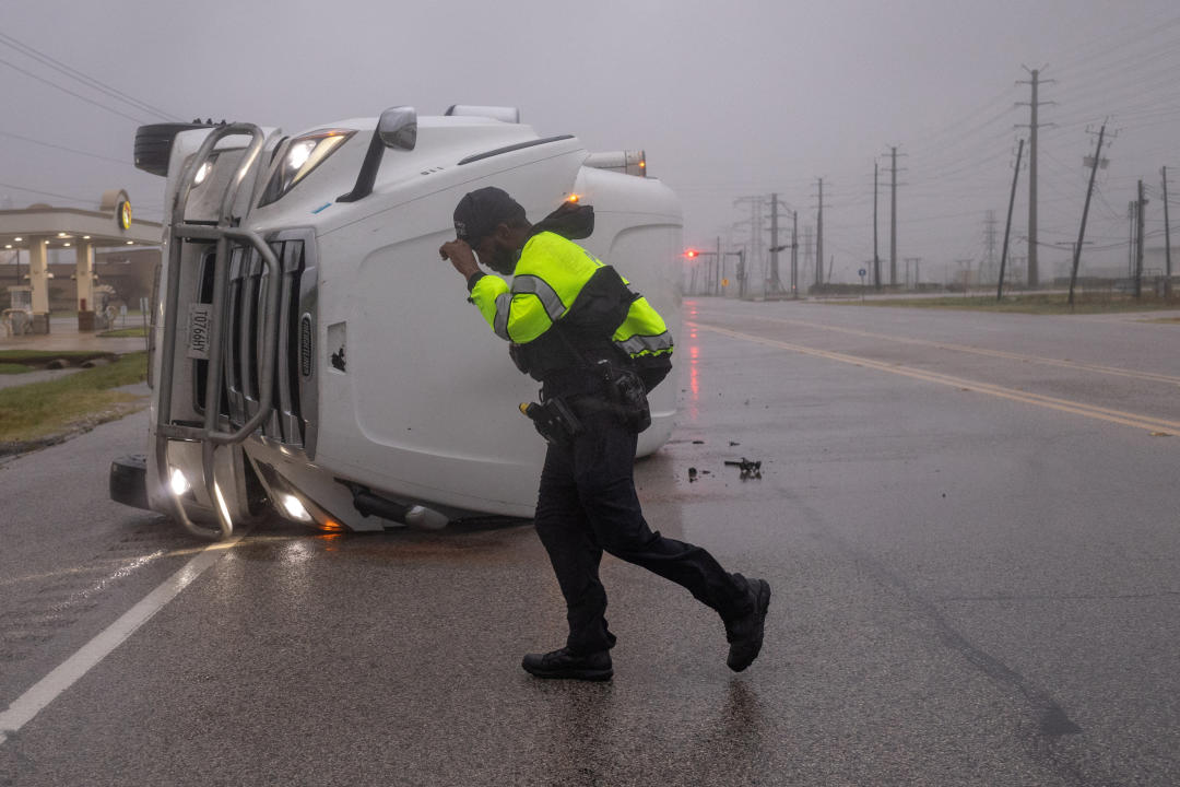 A police officer fights fierce winds as he walks near an overturned semitrailer truck on a desolate road.