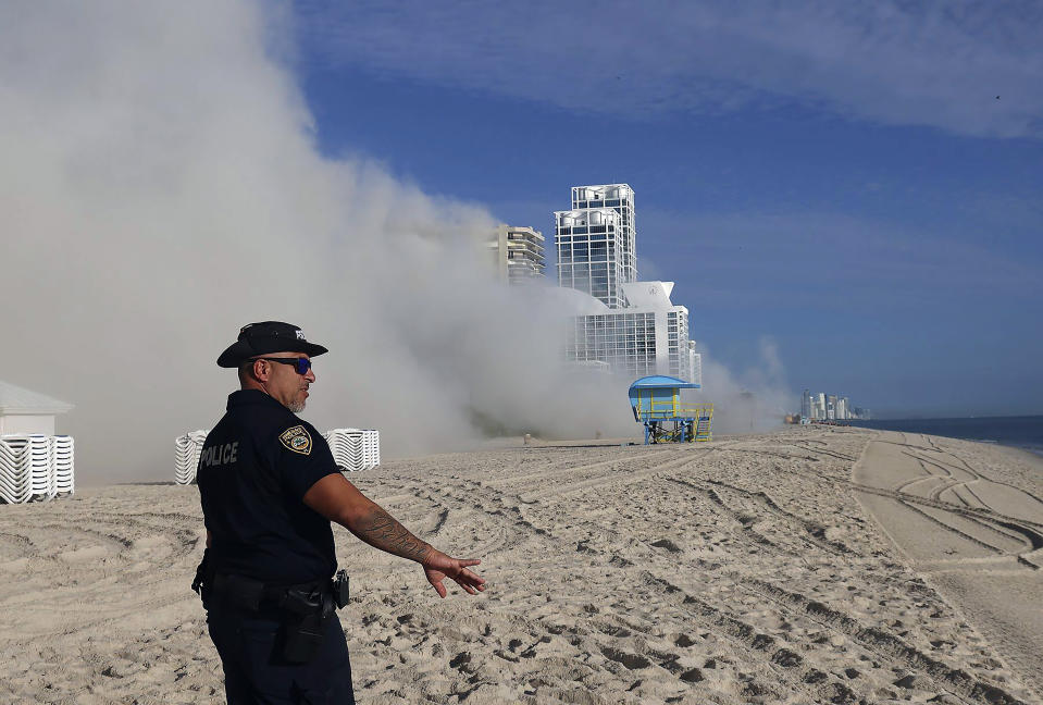 A Miami Beach police officer keeps on-lookers away from a cloud of dust, Sunday, Nov. 13, 2022. The once-luxurious Miami Beach hotel that hosted the Beatles and President John F. Kennedy during its 1960s heyday has been imploded. The 17-story Deauville Hotel fell into itself Sunday after a series of explosions were set off. (Carl Juste/Miami Herald via AP)