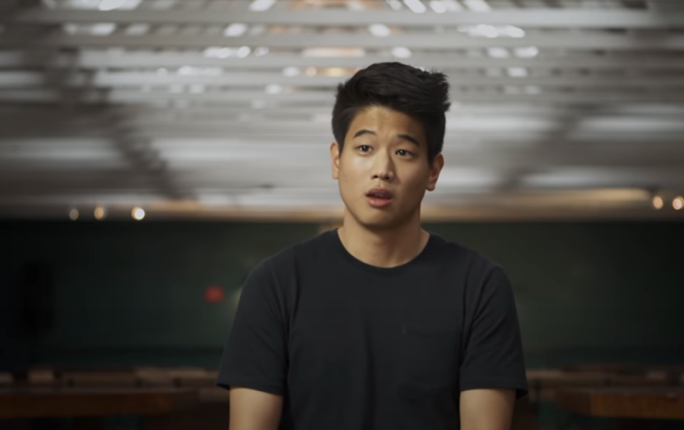 A person with short black hair is sitting indoors, wearing a black T-shirt. The background is blurred with a pattern of ceiling lights