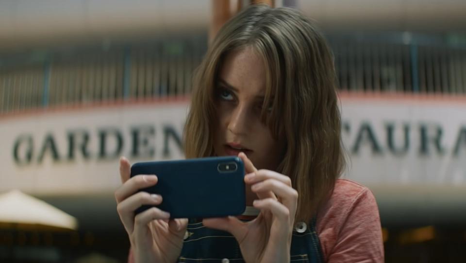 Maya Hawke's Frankie looks cautiously over her phone as she stands before a marquee in a scene from Mainstream.