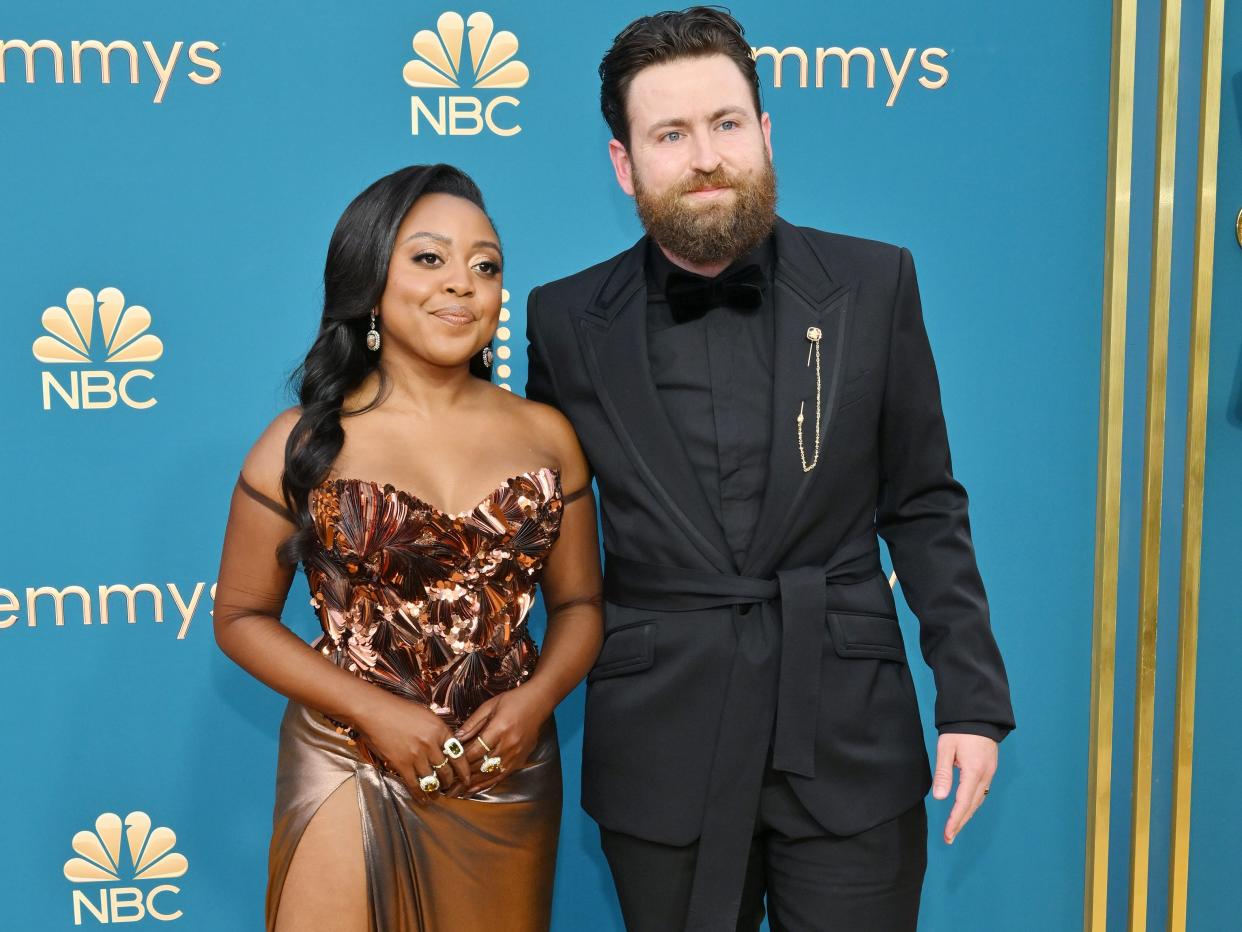 quinta brusnon standing on the gold carpet at the emmys with her hair down, wearing a bronze, strapless dress with a thigh slight. she's standing next to kevin anik, a taller main wearing an all-black suit with a waist tie