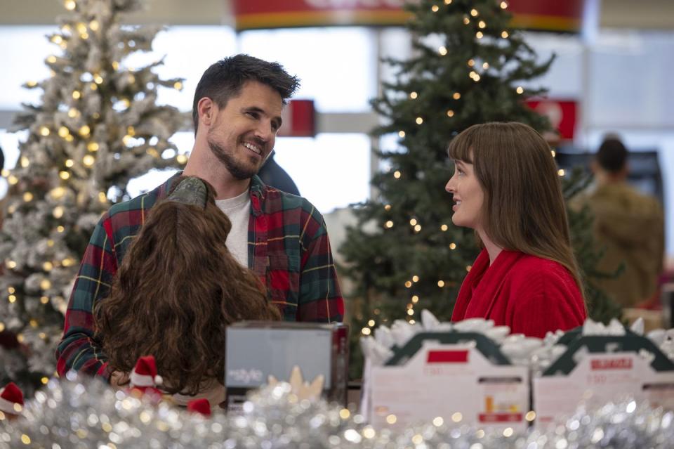 robbie amell and leighton meester star in exmas, a young couple stand in a shop surrounded by christmas decorations and smile at each other