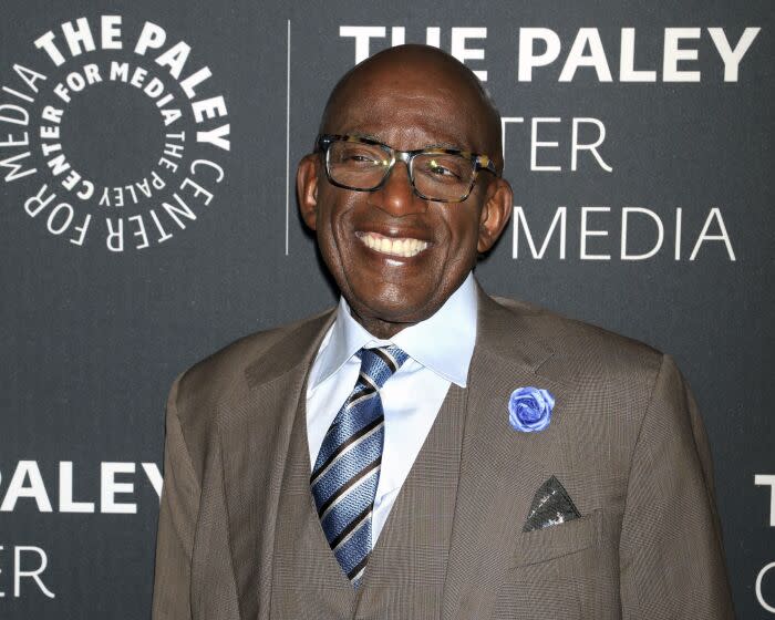 A bald man smiling while wearing glasses, a gray suit with a blue rose on the lapel and a striped blue tie