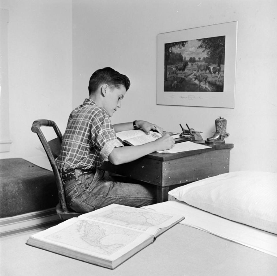 A boy in a plaid shirt is studying at a desk with books and maps spread out. A framed picture hangs above the desk