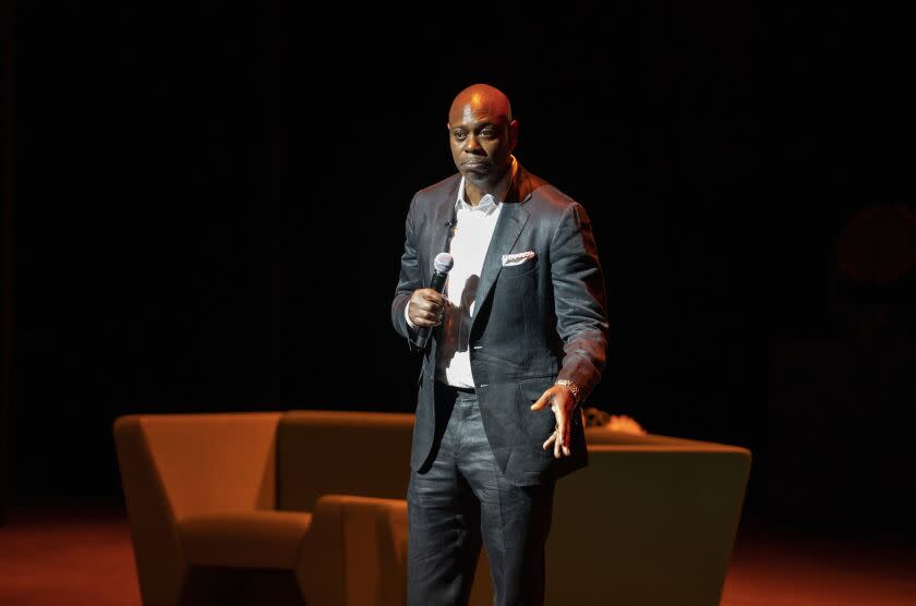 A bald man in a dark suit holding a microphone on a stage with a couch on it