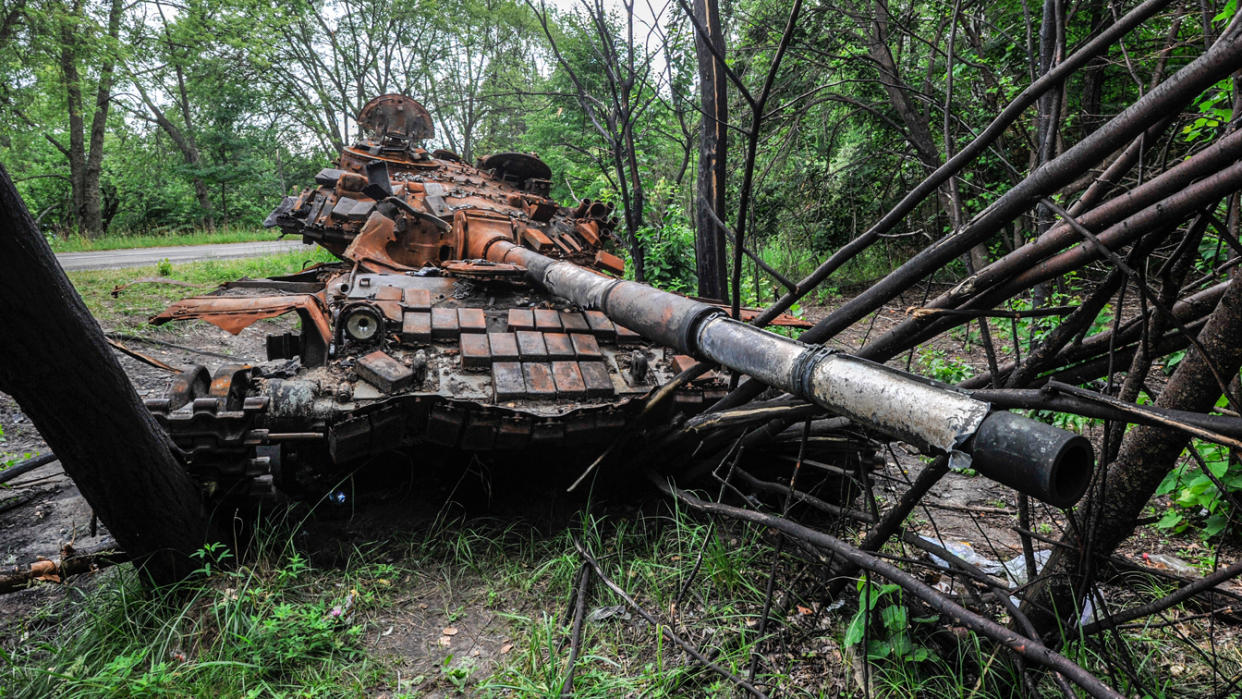 A destroyed tank rests on fallen trees a dozen yards or so from the shoulder of a tree-lined road.