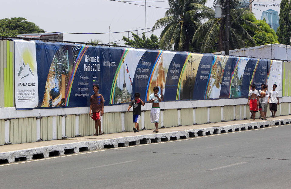 Boys walk past a wall covered with a tarpaulin poster of the ongoing 45th Annual Board of Governors meeting of the Asian Development Bank at suburban Pasay city south of Manila, Philippines, Thursday May 3, 2012. Delegates attending the international conference of the ADB in the Philippines capital may not see what they came to discuss: abject poverty. The makeshift, temporary wall on both sides of the bridge from the airport to downtown Manila, hides a sprawling slum along a garbage-strewn creek in the background. (AP Photo/Bullit Marquez)